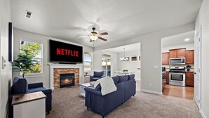 Una sala de estar moderna con chimenea, un televisor grande que muestra 'NETFLIX' y una cocina visible a través de un vano abierto.