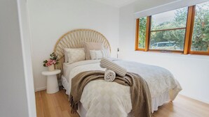 A bedroom with a round wooden headboard, a bedside table with a vase of flowers, a large window with a view of greenery, and a bed with a quilt and a throw blanket. - Chic Beachside Stay – Near River, Shops & Cafés (Ocean Grove)
