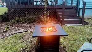 A square fire pit with flames, surrounded by grass and mulch, with a wooden deck and stairs in the background. - Village District - Historic 2-bedroom house in Raleigh (Raleigh)