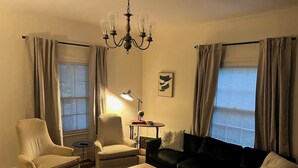 A living room with a striped armchair, a black sofa, a wooden coffee table with a book and a cup, a floor lamp, and a chandelier. - Village District - Historic 2-bedroom house in Raleigh (Raleigh)