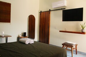 A hotel room with a bed, a wooden chair, a small table, a TV mounted on the wall, and a shelf with a plant. - Casa de Alicia - Xaguar Room (Oaxaca)