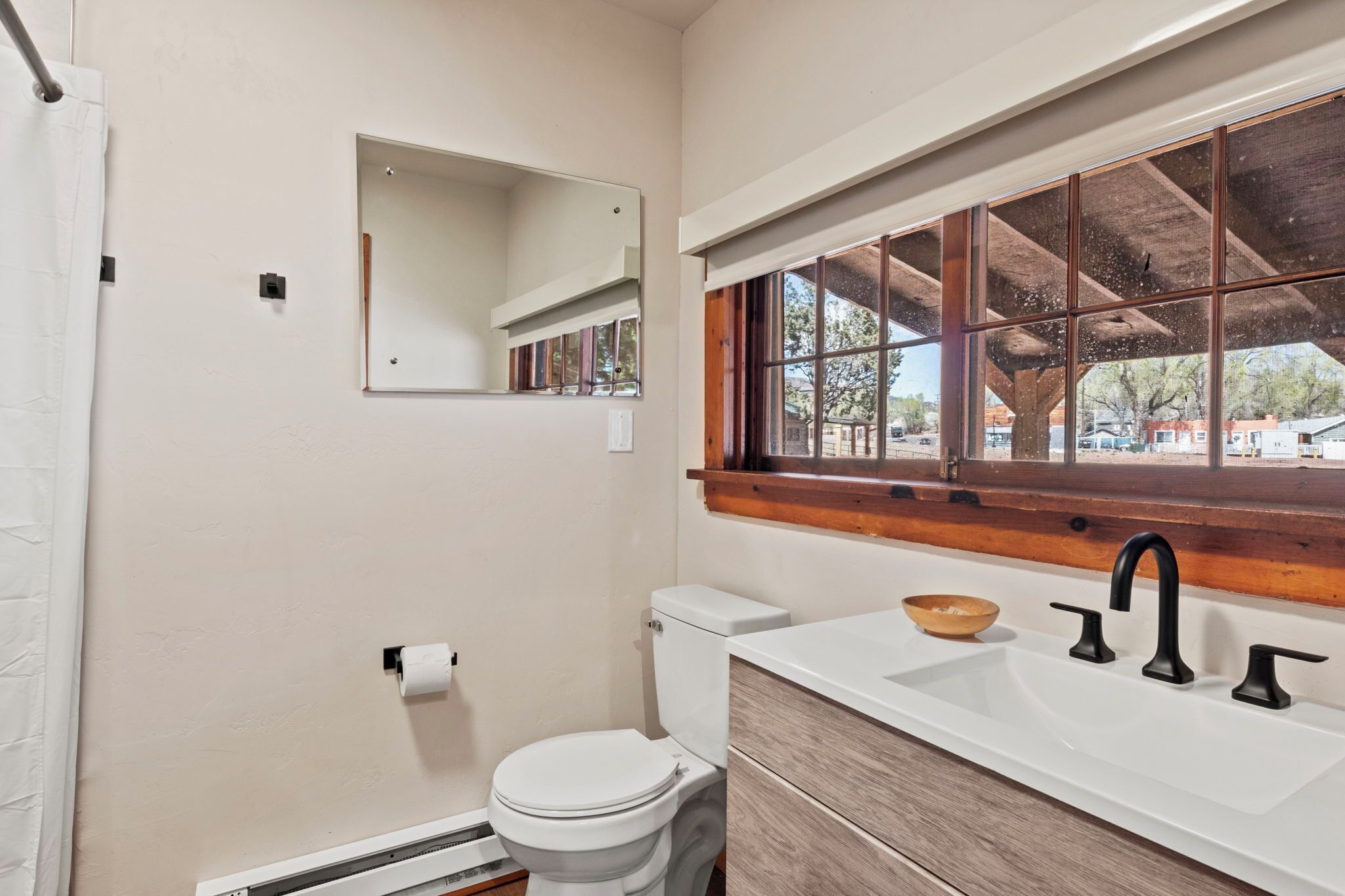 Un baño con inodoro blanco, tocador de madera con lavamanos, espejo y ventana con vista al exterior.