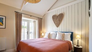 A bedroom with a bed, a heart-shaped wall decoration, a lamp, and a window with curtains. - Cowlands 3 bed, barn conversion, pet friendly, three bedrooms, Sleeps 6 (Truro)