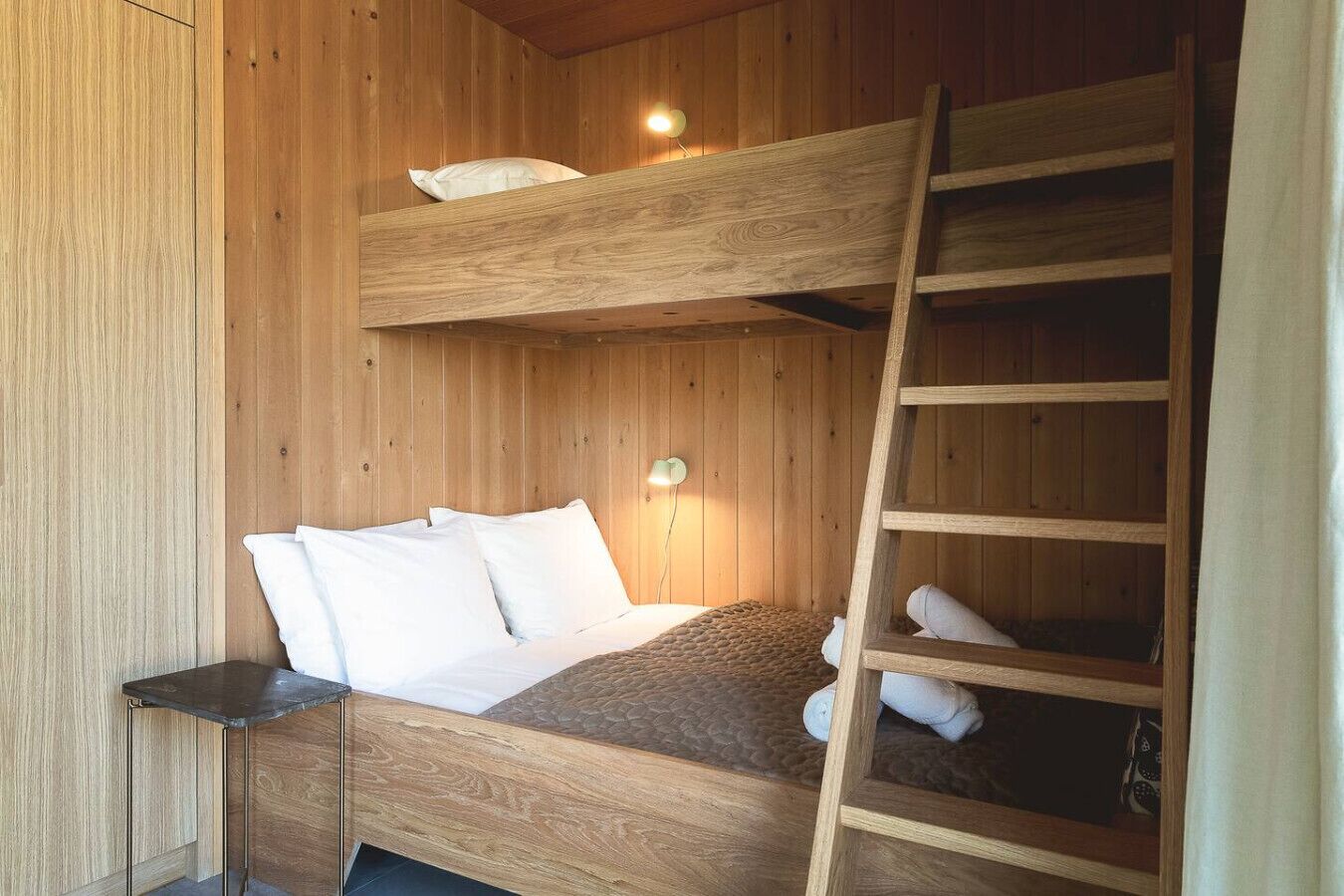 A bunk bed room with a wooden ladder, a bedside lamp, and a small table.