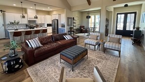A spacious living area with a leather sofa, a patterned ottoman, and a plaid armchair. A kitchen with white cabinets and a bar area is visible in the background.
