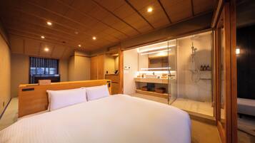 A hotel room with a large bed, a wooden ceiling, a bathroom with a glass shower enclosure, and a small desk area.