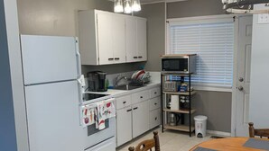 A compact kitchen with white cabinets, a refrigerator, a microwave, and a sink. There is a dining table with chairs and a window with blinds.