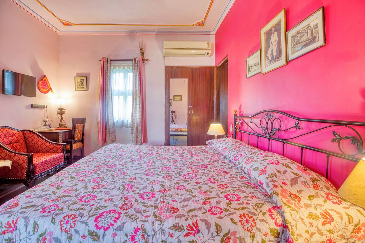 Une chambre d’hôtel avec une couvre-lit à motifs floraux, un fauteuil rouge, un bureau avec une lampe et un téléviseur fixé au mur.