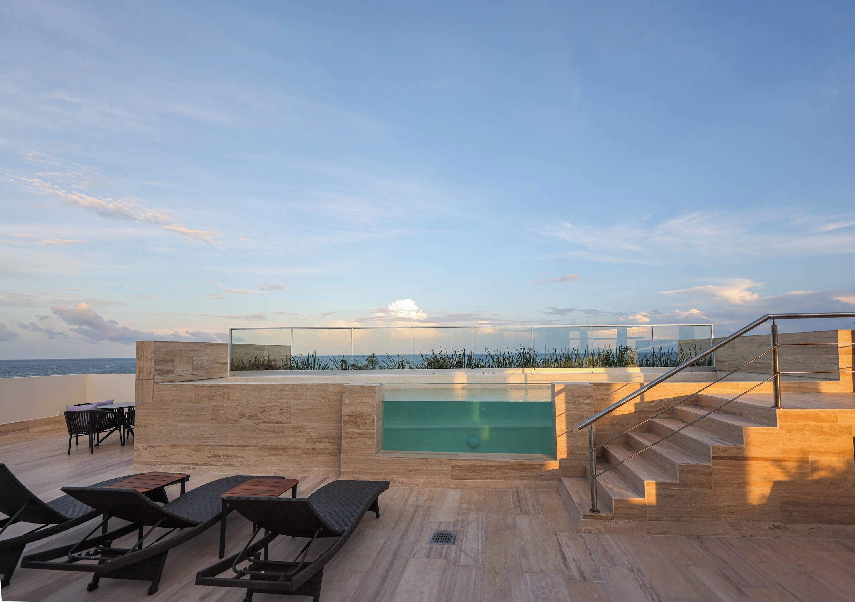 Une terrasse sur le toit dotée d’une piscine à débordement à ciel ouvert, de chaises longues et d’un escalier menant à un niveau supérieur.