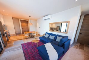 A modern living room with a blue sofa, a wooden dining table, and a kitchen area in the background. - Distrito Puerto – Ocean & Mangrove Views (Puerto Morelos)