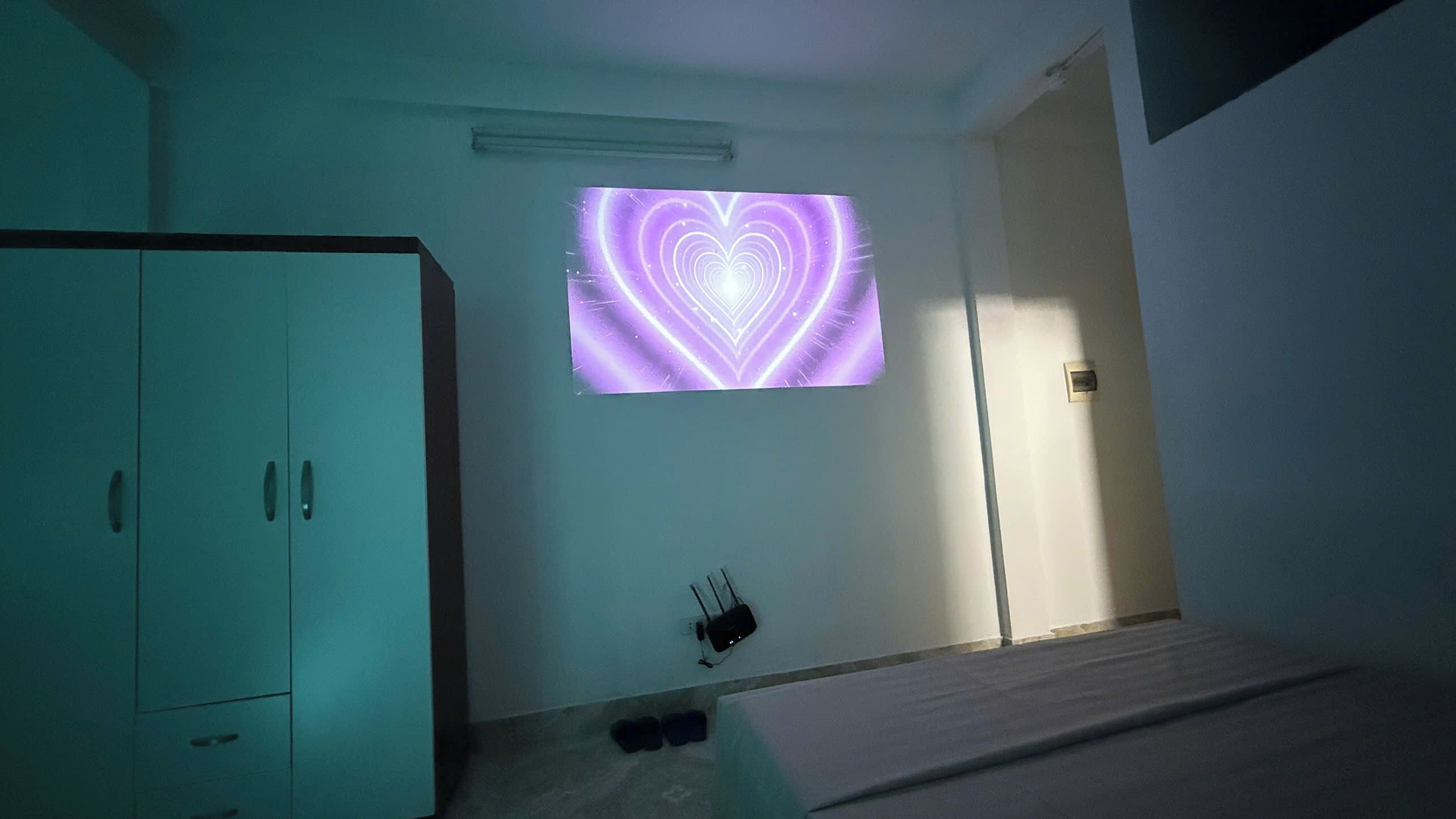 Ein Zimmer mit einem Bett, einem Kleiderschrank, einem an der Wand befestigten Projektor, der ein Herzmuster zeigt, und einer Steckdose mit eingestecktem Gerät.