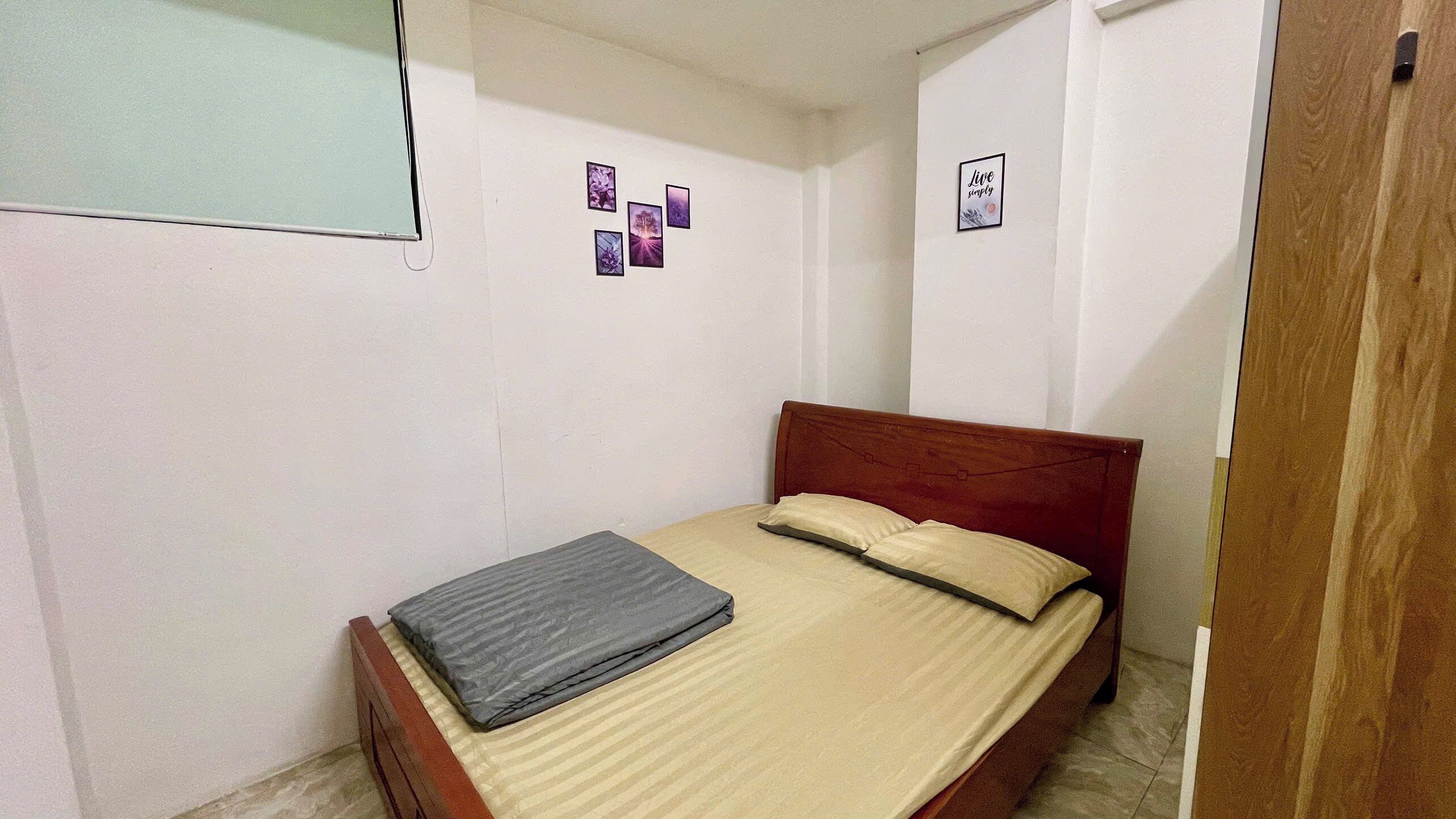 A small, single-bed room with a wooden headboard, a beige bedspread, and a grey pillow. There are three small pictures on the wall and a 'Live simply' sign.