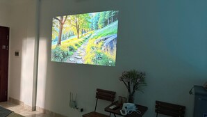 Une pièce avec un grand téléviseur fixé au mur, diffusant une scène de nature, un banc en bois, une petite table avec un vase de fleurs et une prise électrique au mur.