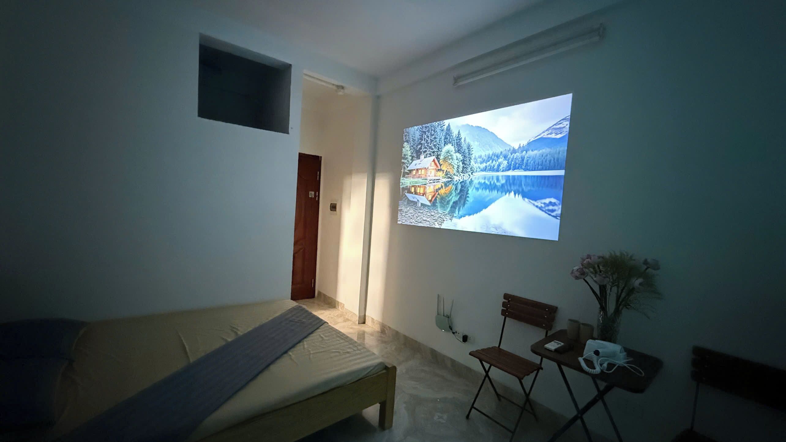 Ein Schlafzimmer mit einem Bett, einem kleinen Schreibtisch, einem Stuhl und einem an der Wand montierten Fernseher, der ein Landschaftsbild zeigt.