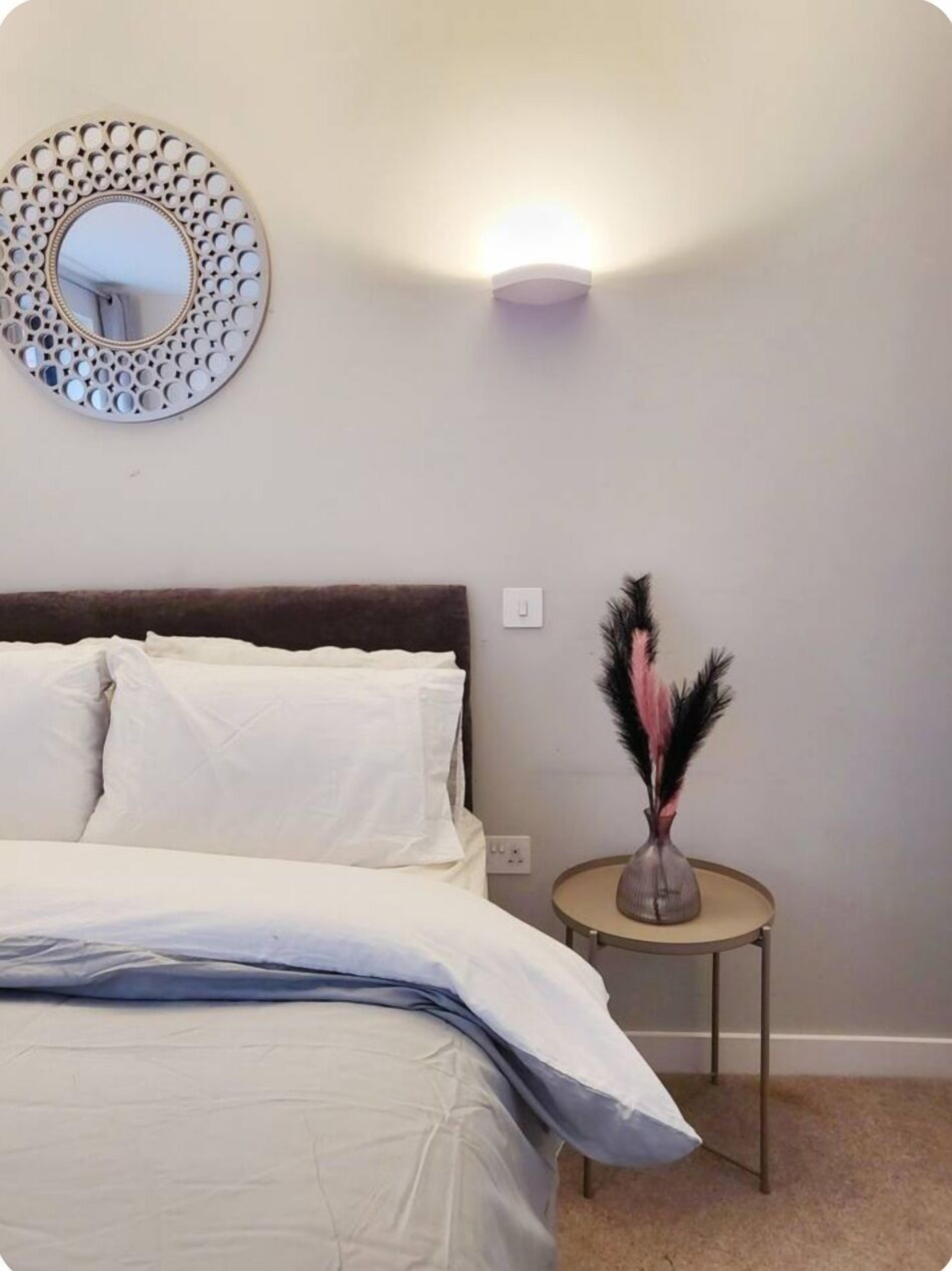 A bedroom with a bed, a round mirror, a wall-mounted light, and a small table with a vase.