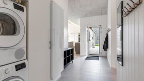 A laundry room with stacked washers and dryers, a black storage unit, and a hallway leading to a kitchen area. - VillaVilla 806 - Fjellerup, Østjylland (Glesborg)