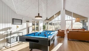 Una mesa de billar en una habitación luminosa con ventanas amplias, un sofá y una mesa con un jarrón.