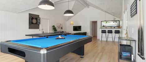 Una mesa de billar en una habitación con techo alto y paredes blancas. Hay dos lámparas colgantes sobre la mesa.