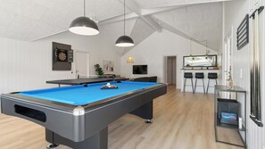 Ein Billardtisch in einem Raum mit hoher Decke und weißen Wänden. Zwei Hängelampen sind über dem Tisch angebracht.