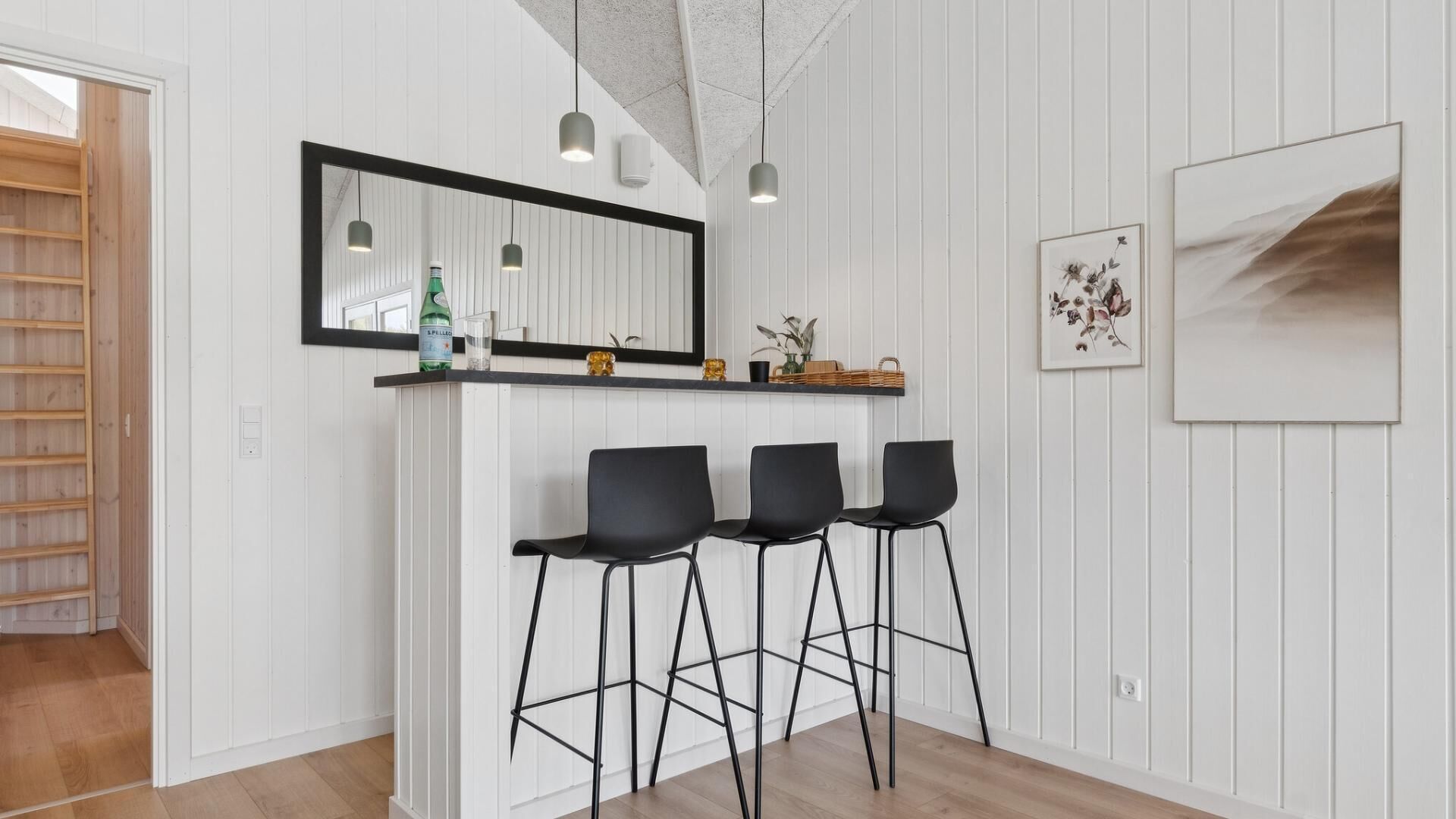 A modern kitchen with white wooden paneling, a black-framed mirror, pendant lights, and three black bar stools.