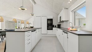 Une cuisine moderne avec des meubles blancs, un îlot central, des luminaires suspendus et une vue sur l’extérieur par une fenêtre.