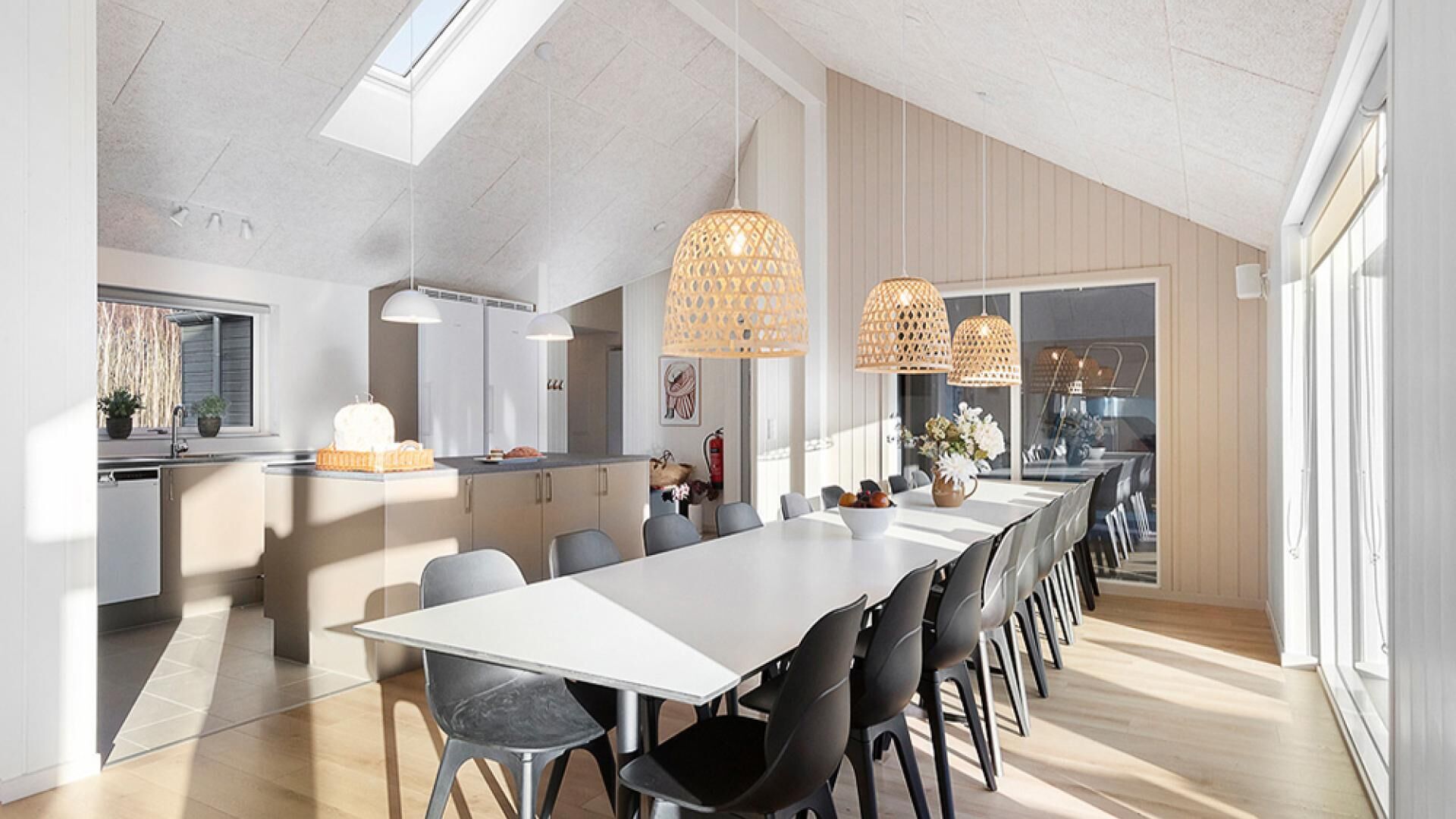 A modern kitchen with a long dining table, pendant lights, and a view of the outdoors.