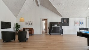 A spacious living room with a high ceiling, wooden flooring, a brown sofa, a green armchair, a TV, a ping pong table, and a dartboard.