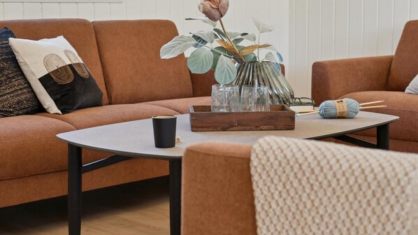 A living room with a brown sofa, a coffee table, a vase with a flower, a knitting ball, and a pair of glasses.