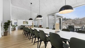 A modern dining area with a long table, chairs, pendant lights, and a view of the outdoors.