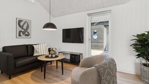 A modern living room with a black sofa, a round coffee table, a grey armchair, a TV mounted on the wall, and a large window with a view of the outside.