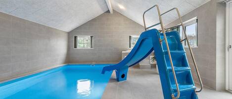 An indoor swimming pool with a blue slide, a ladder, and two windows.