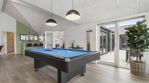 Une table de billard dans une pièce dotée d’un plancher en bois, d’un luminaire suspendu et d’une porte-fenêtre coulissante donnant sur l’extérieur.