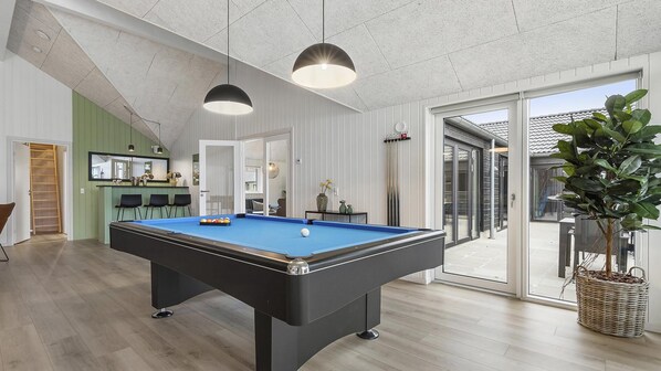 Une table de billard dans une pièce dotée d’un plancher en bois, d’un luminaire suspendu et d’une porte-fenêtre coulissante donnant sur l’extérieur.