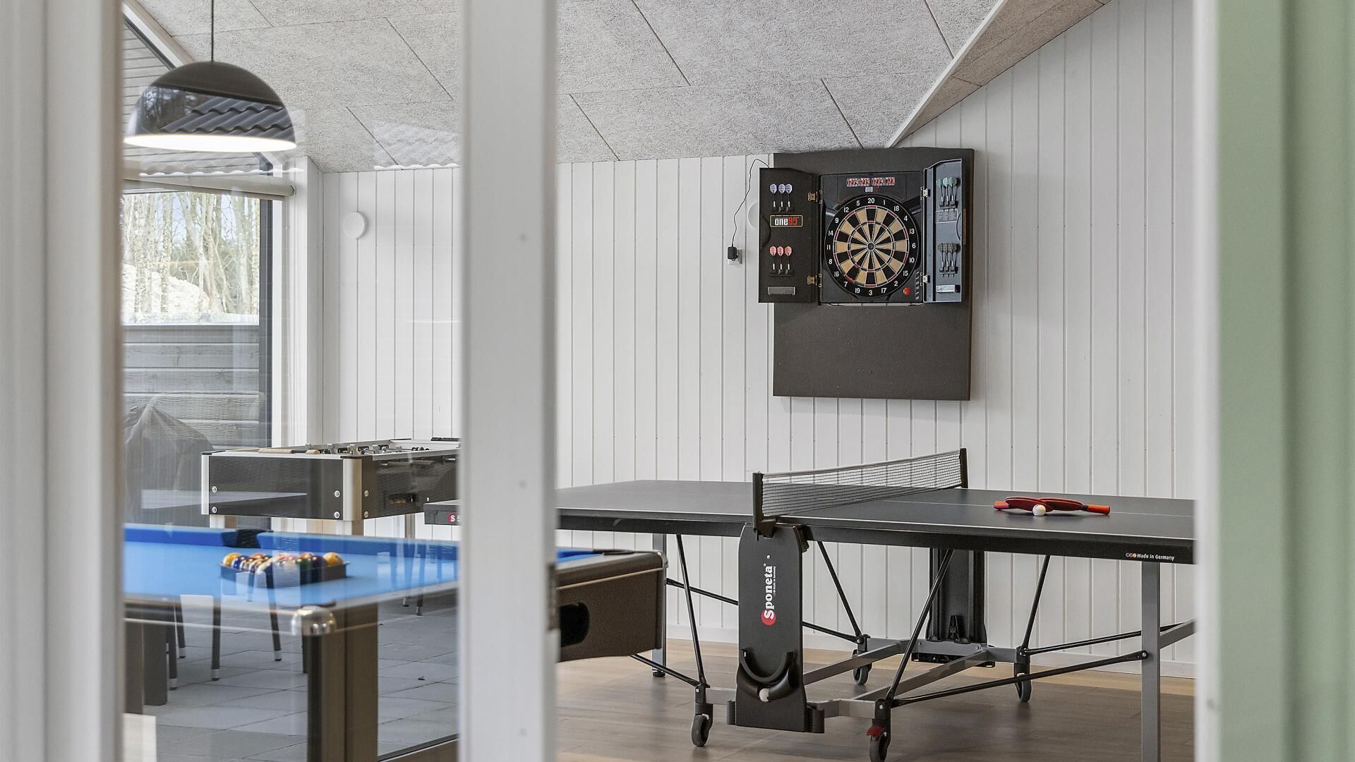 A billiards table, a dartboard, and a ping pong table in a room with white walls and a visible window.