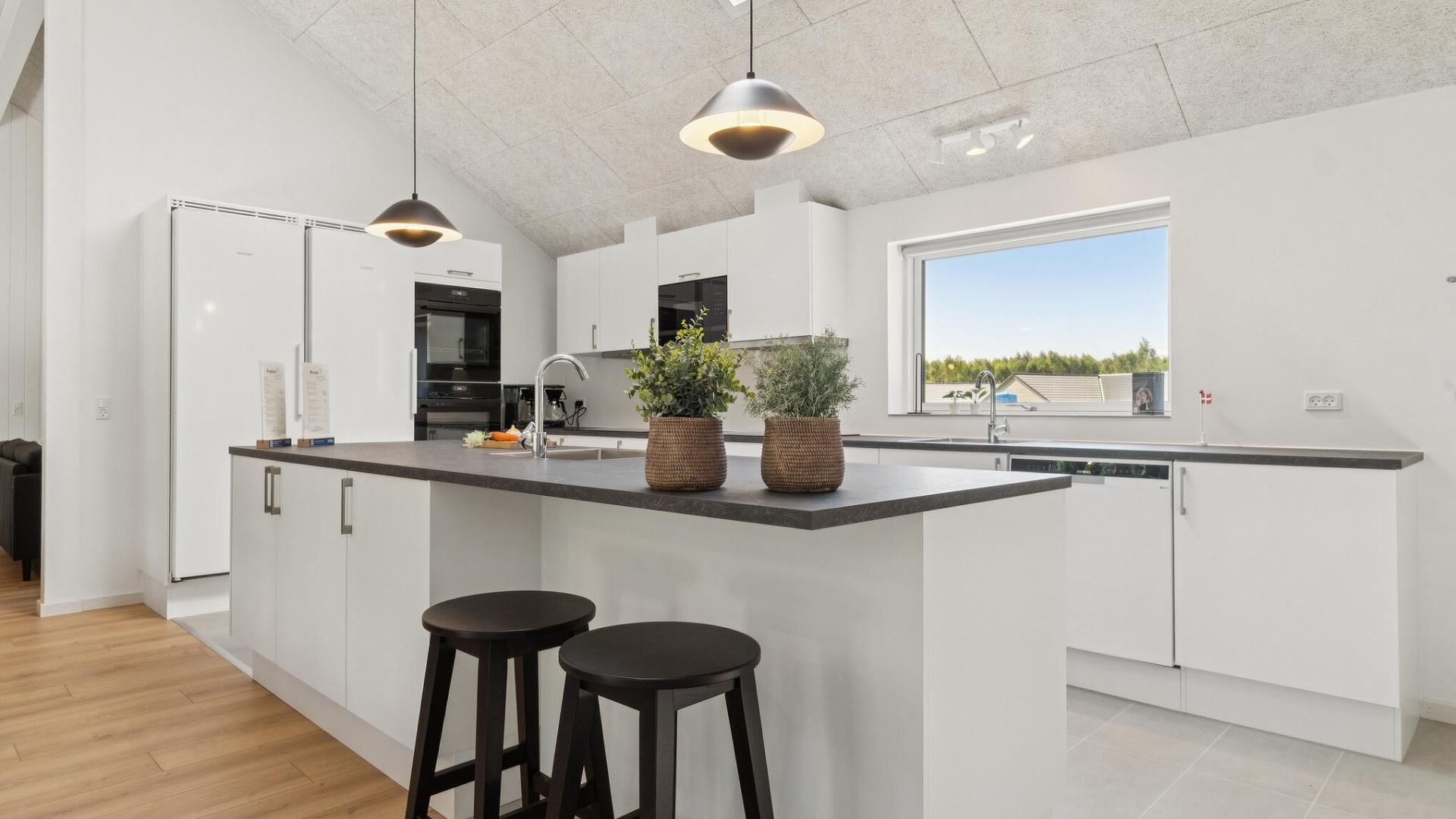 Una cocina moderna con armarios blancos, una isla grande con taburetes, iluminación colgante y vistas al exterior a través de una ventana.