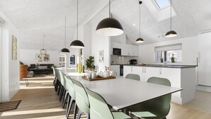 Een ruime eethoek met een lange tafel, groene stoelen, hanglampen en uitzicht op de keuken.
