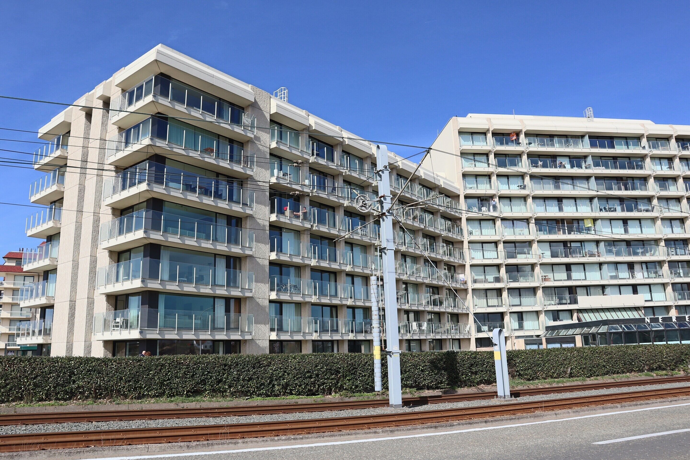 A multi-story hotel with balconies, a hedge in front, and a railway track nearby.