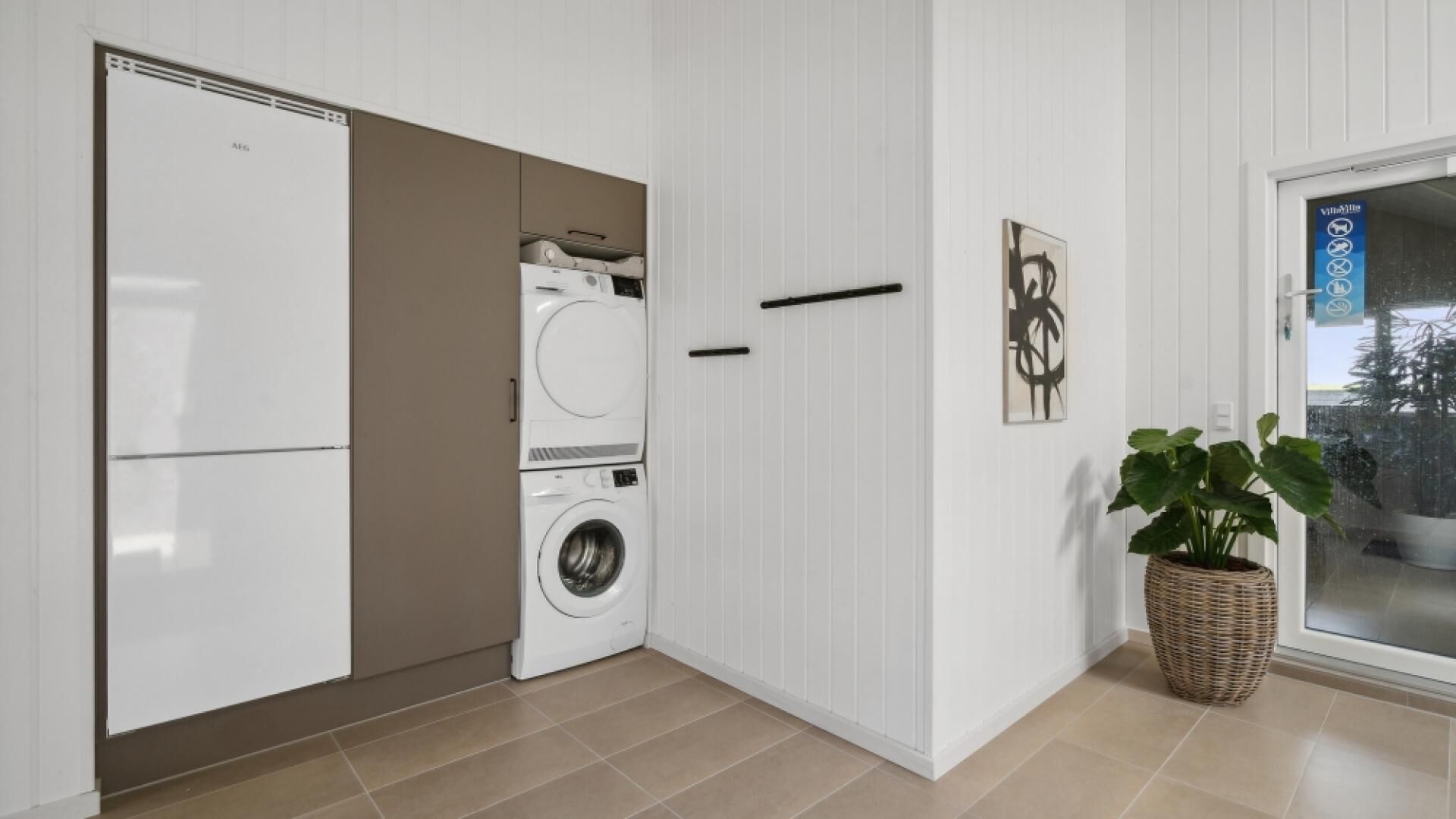 Une petite buanderie comprenant un lave-linge et un sèche-linge, une porte avec une poignée et une plante en pot.