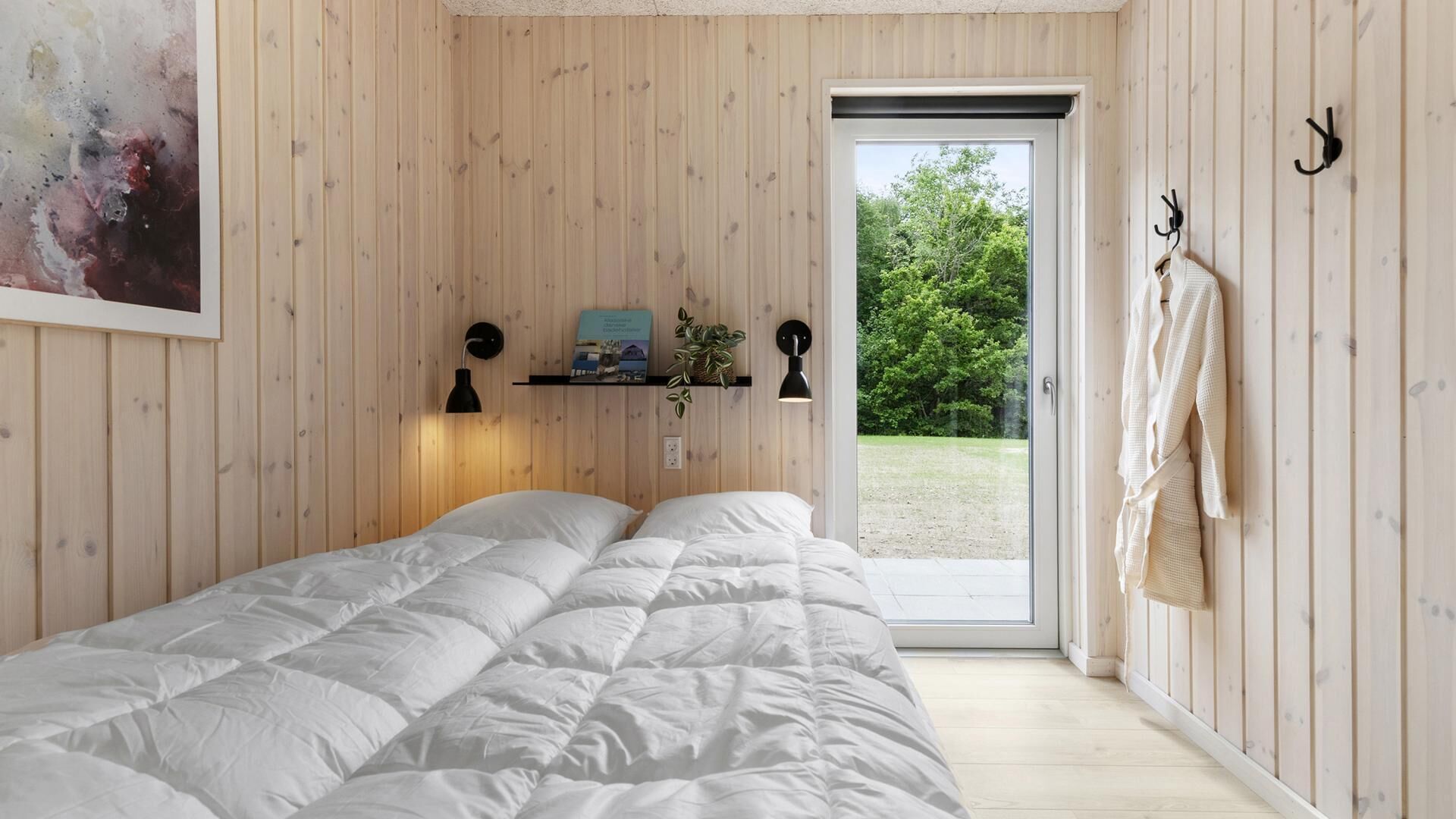Une chambre à coucher avec un lit, une étagère ornée d’une plante, un tableau et une porte donnant sur l’extérieur.