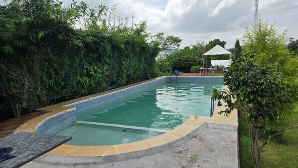 En swimmingpool med en kant som är klädd i kakel, omgiven av grönska och med en vit, pergola-liknande konstruktion i bakgrunden.