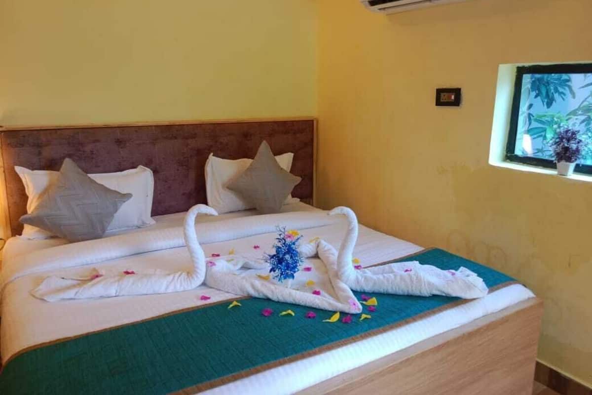 A hotel room with a bed featuring a swan-shaped towel arrangement, a green bedspread, and a window with a plant view.