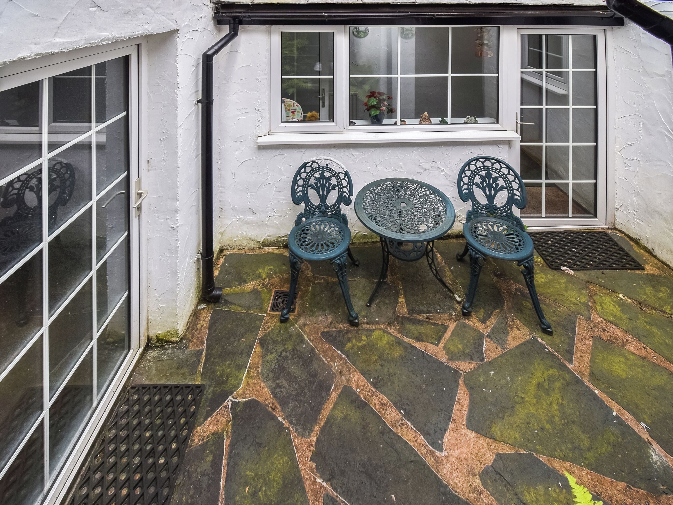 Eine Terrasse mit Steinboden, zwei verzierten Metallstühlen, einem kleinen runden Tisch und einem Fenster mit Pflanze auf dem Fensterbrett.