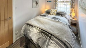 Un dormitorio con cama, mesita de noche, lámpara, ventana con cortinas y puerta.