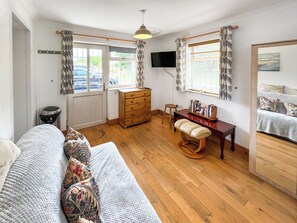 A cozy room with a wooden floor, a sofa with patterned cushions, a desk with a chair, a television, and a window with curtains.