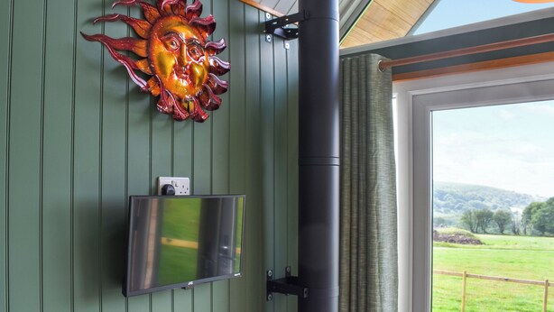 Ein Zimmer mit grüner Wand, einem an der Wand montierten Fernseher, Wandkunst, die einem Sonnenmotiv ähnelt, und einem großen Fenster mit Blick auf ein grünes Feld und Hügel.