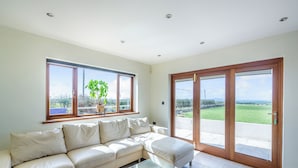 Ein geräumiges Wohnzimmer mit einem beigefarbenen Sofa, einem Glastisch und Blick durch große Schiebetüren in einen Garten.
