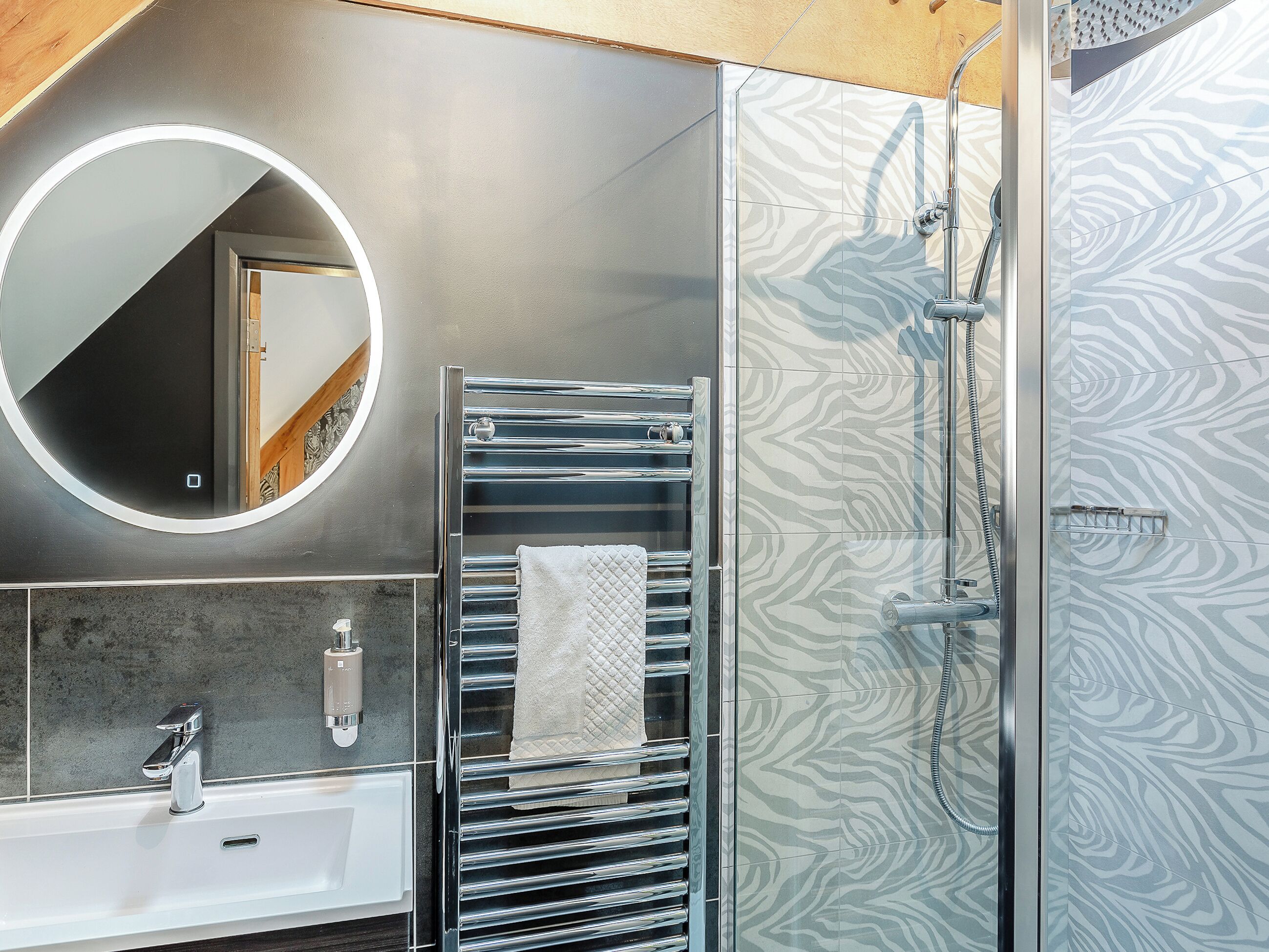 A modern bathroom with a round mirror, a towel rack, a shower with a patterned wall, and a sink with a soap dispenser.