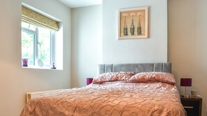 A bedroom with a bed, a window with curtains, a bedside table with a lamp, and a framed picture on the wall.