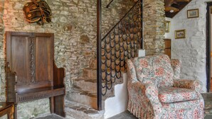 Une pièce rustique avec un mur en pierre, une armoire en bois, un fauteuil à motifs floraux et un escalier doté d’une rampe en métal.