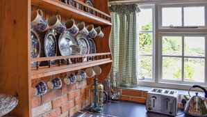 Une cuisine avec des étagères en bois où sont rangées des assiettes en céramique bleue et blanche, un mur en briques, une fenêtre donnant sur un paysage verdoyant et un grille-pain posé sur le plan de travail.